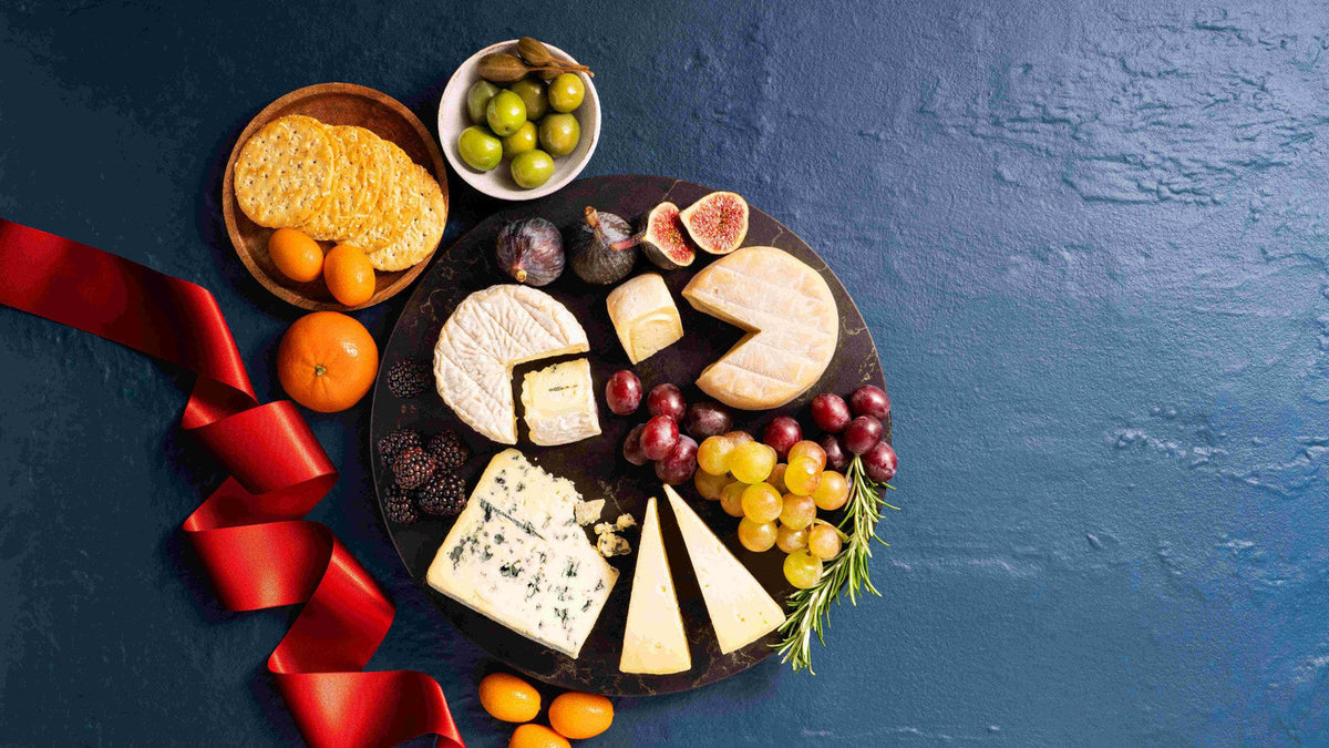 A Christmas cheeseboard of Caws Cenarth Welsh cheeses — Perl Las Deli, Perl Las, Golden Cenarth and Thelma's Traditional Welsh Caerffili — with grapes, figs, olives, crackers and a red ribbon on a dark blue background