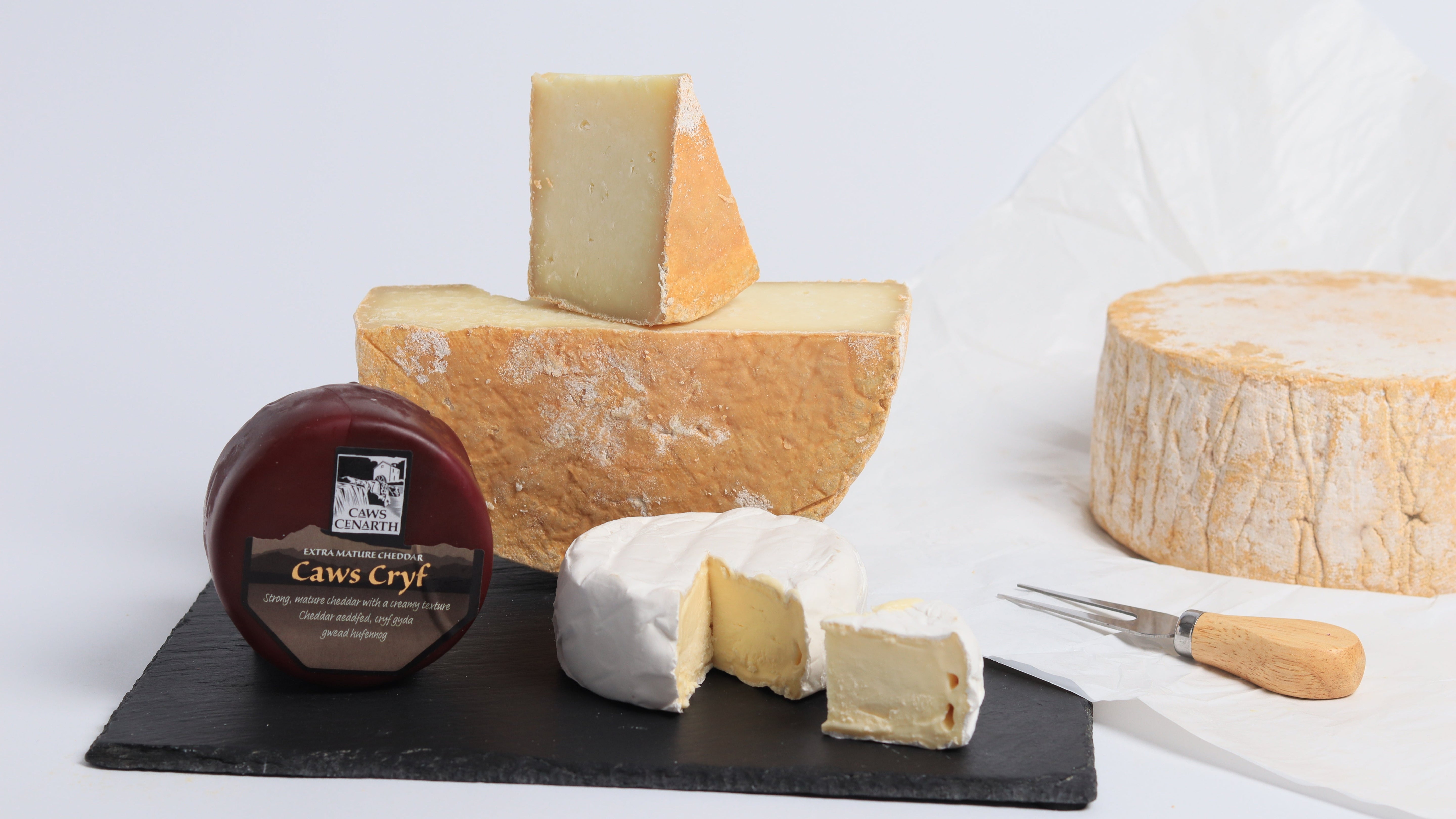 3 pieces of artisan cheeses with a wheel of cheese and cheese fork in the background