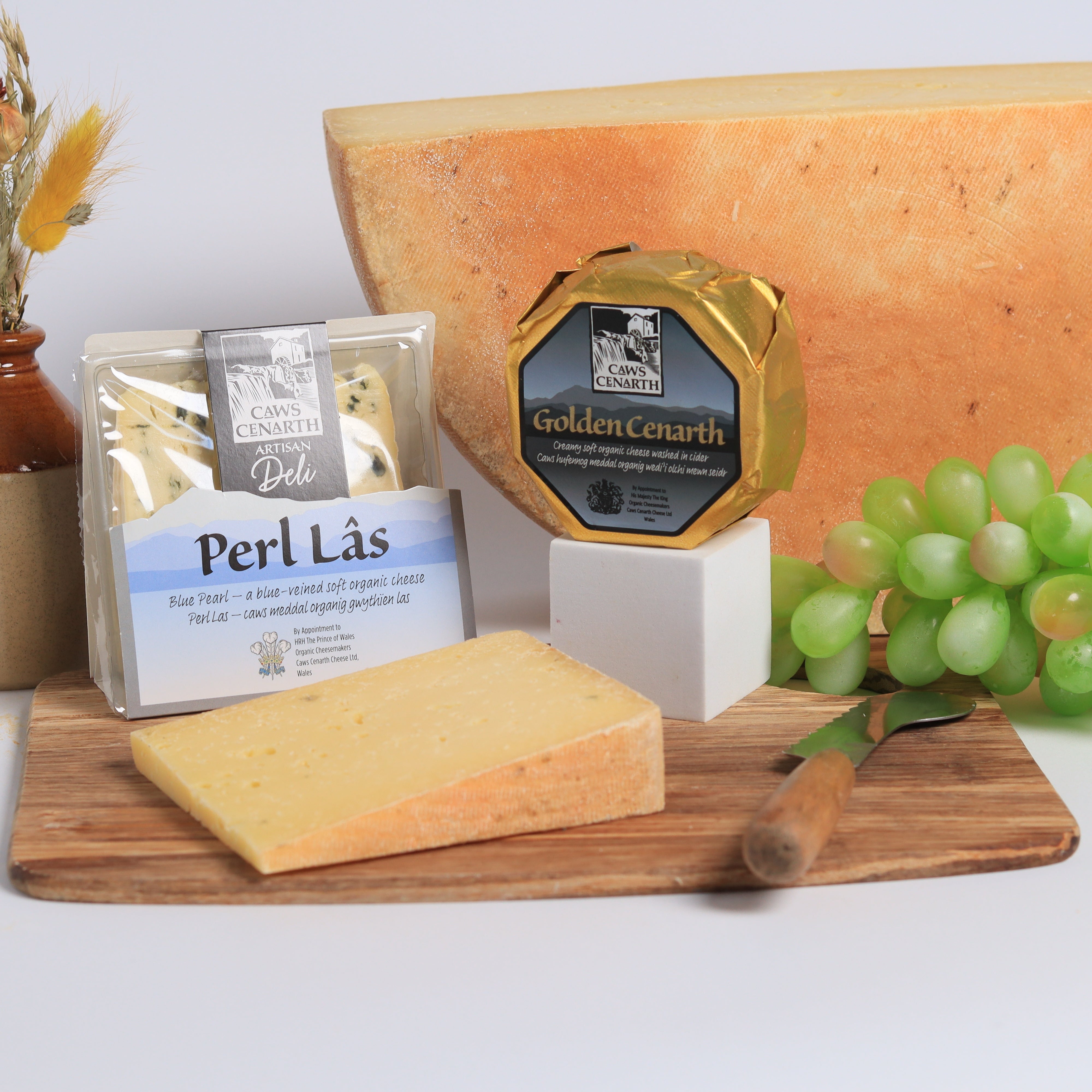 Assorted cheeses on a wooden board with a vase and grapes on a white background